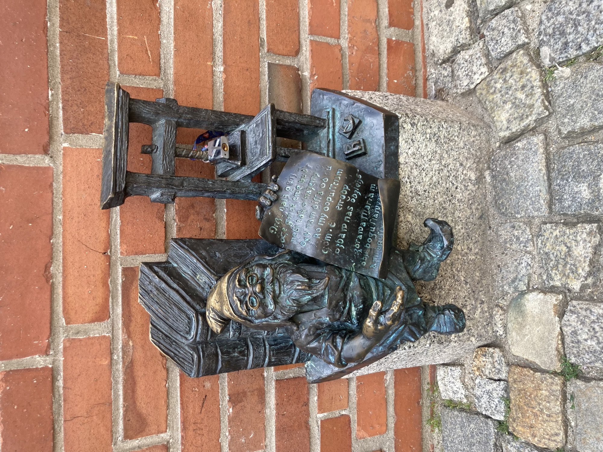 Metallstatue eines Zwergs neben einer Druckerpresse, der Zwerg hat drei Bücher hinter seinem Rücken und zeigt ein ausgerolltes Blatt Papier