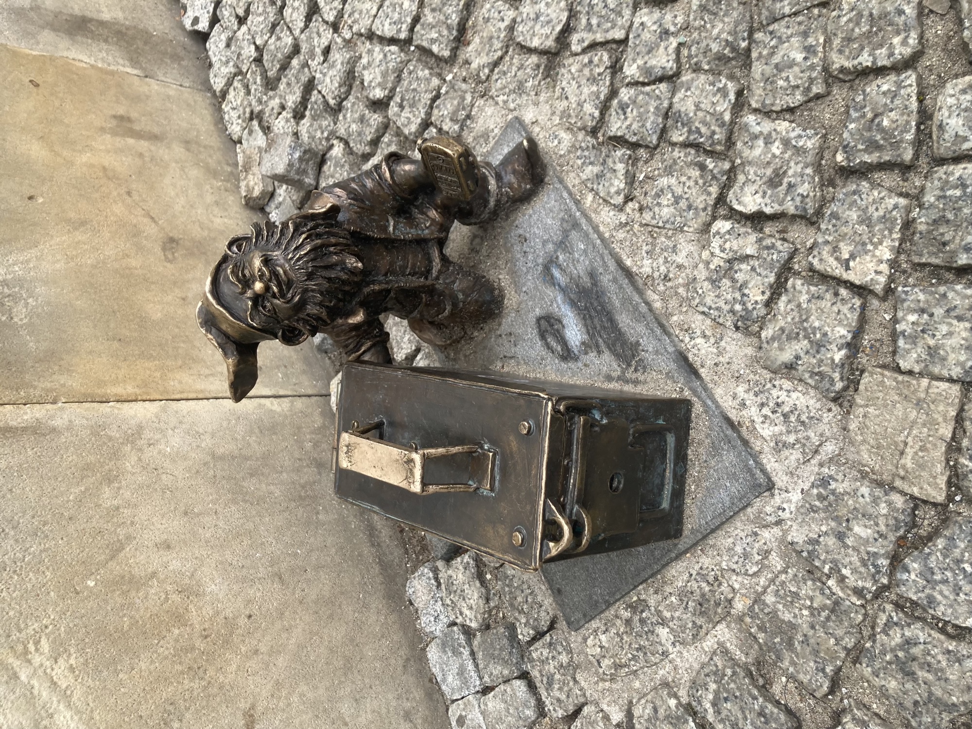 Metallstatue eines Zwergs mit einer Metallkiste mit einem Griff obendrauf, der Zwerg hält ein GPS-Gerät in der Hand