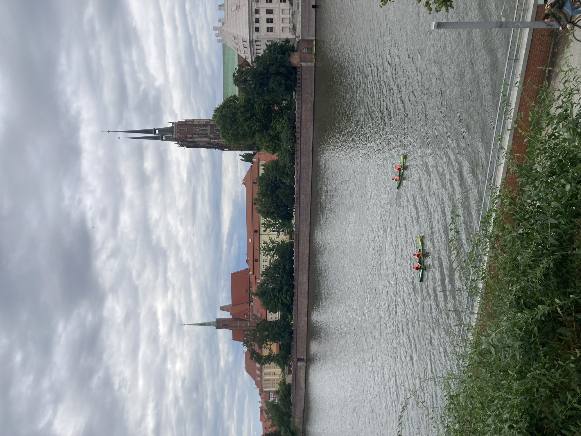 auf einem Fluss zwei Kajaks mit jeweils zwei Personen in Warnwesten, dahinter am anderen Ufer sind drei Kirchtürme zu sehen
