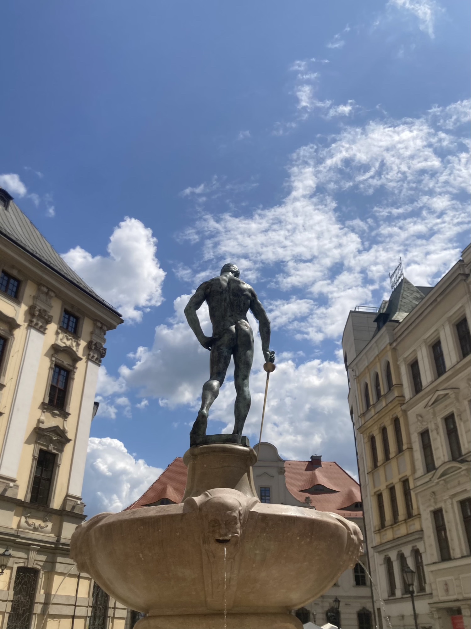 Brunnen zwischen zwei Häusern, auf dem Brunnen steht eine nackte männliche Statue mit dem Rücken zur Kamera, mit der rechten Hand auf einen Säbel, die linke Hand in die Hüfte gestützt, dahinter blauer Himmel mit kleinen Wölkchen