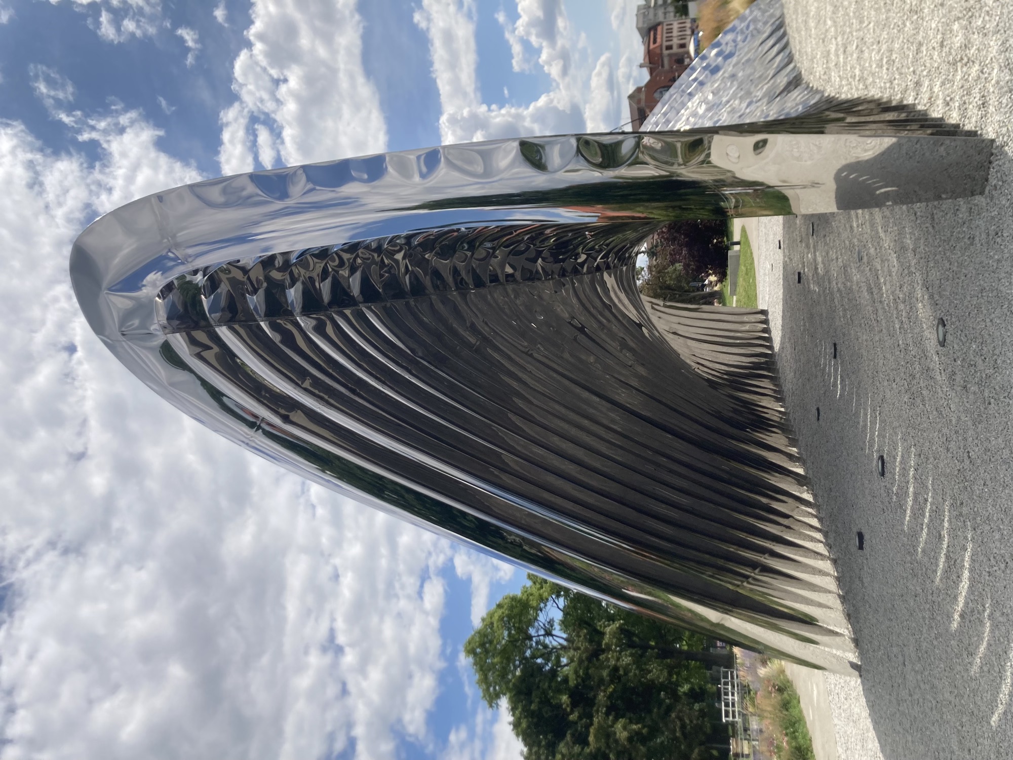 metallisch glänzende Skulptur unter leicht bewölktem blauen Himmel, die Skulptur sieht aus wie eine Art Welle, in der Mitte können Besucher:innen durchgehen