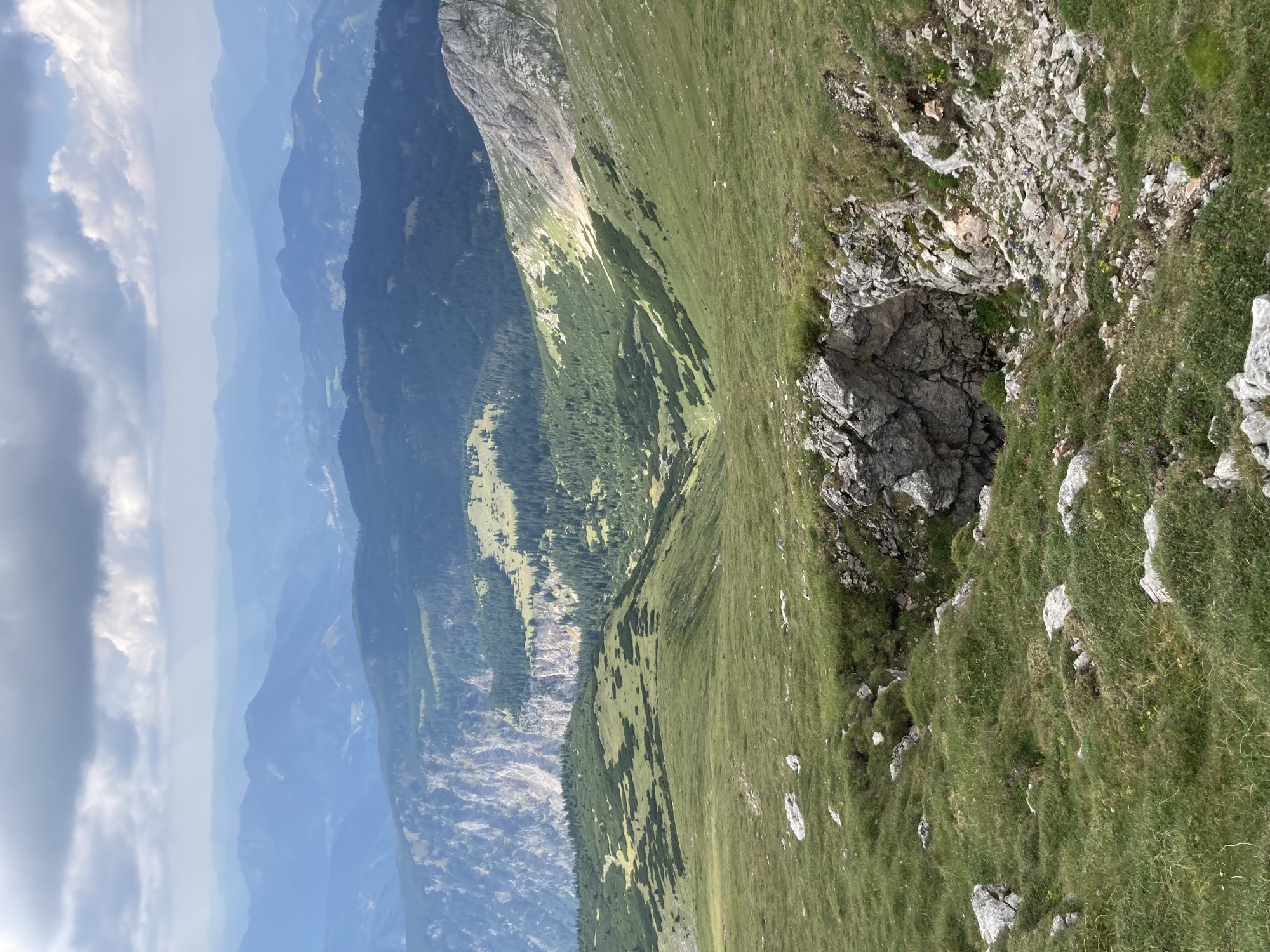im Vordergrund ein Steinloch im Boden, im Hintergrund Bergpanorama mit weithin dunkel bewaldeten Hügeln und flauschigen Wolken