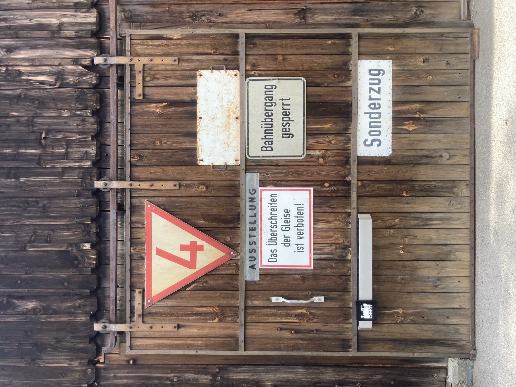 geschlossenes Tor zu einer hölzernen Scheune, außen am Tor mehrere Schilder: ein weißes Dreieck mit rotem Rand, das auf der Spitze steht, darin die Zahl 4, rechteckige Schilder mit den Texten „Ausstellung“, „Bahnübergang gesperrt“, „Sonderzug“, „Das Überschreiten der Gleise ist verboten“, am Türgriff ist eine kleine Dampflokomotive erkennbar