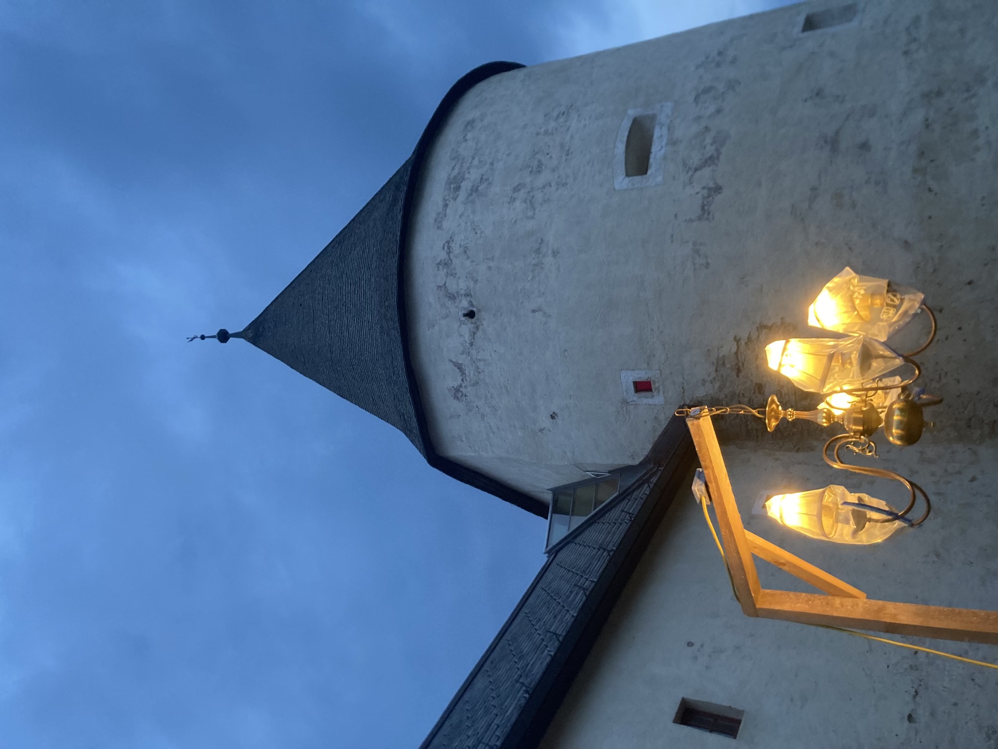 massiver runder Turm mit einem spitzen Dach vor bewölktem Himmel, das Licht ist Abenddämmerung, im Vordergrund unten ein Leuchter mit fünf Lampen an einer Art Holzgalgen, die einzelnen Lampen sind in Plastiksäcken verpackt