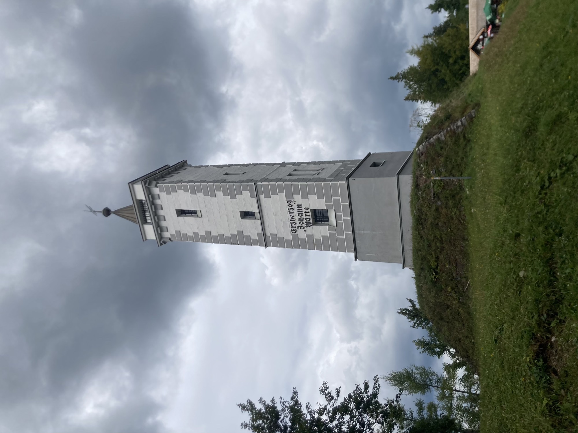 grau-weiß gestrichener Aussichtsturm mit der Aufschrift Erzherzog Johann Warte, gekrönt von einer Plattform und einem spitzen Turm vor einem düster bewölkten Himmel